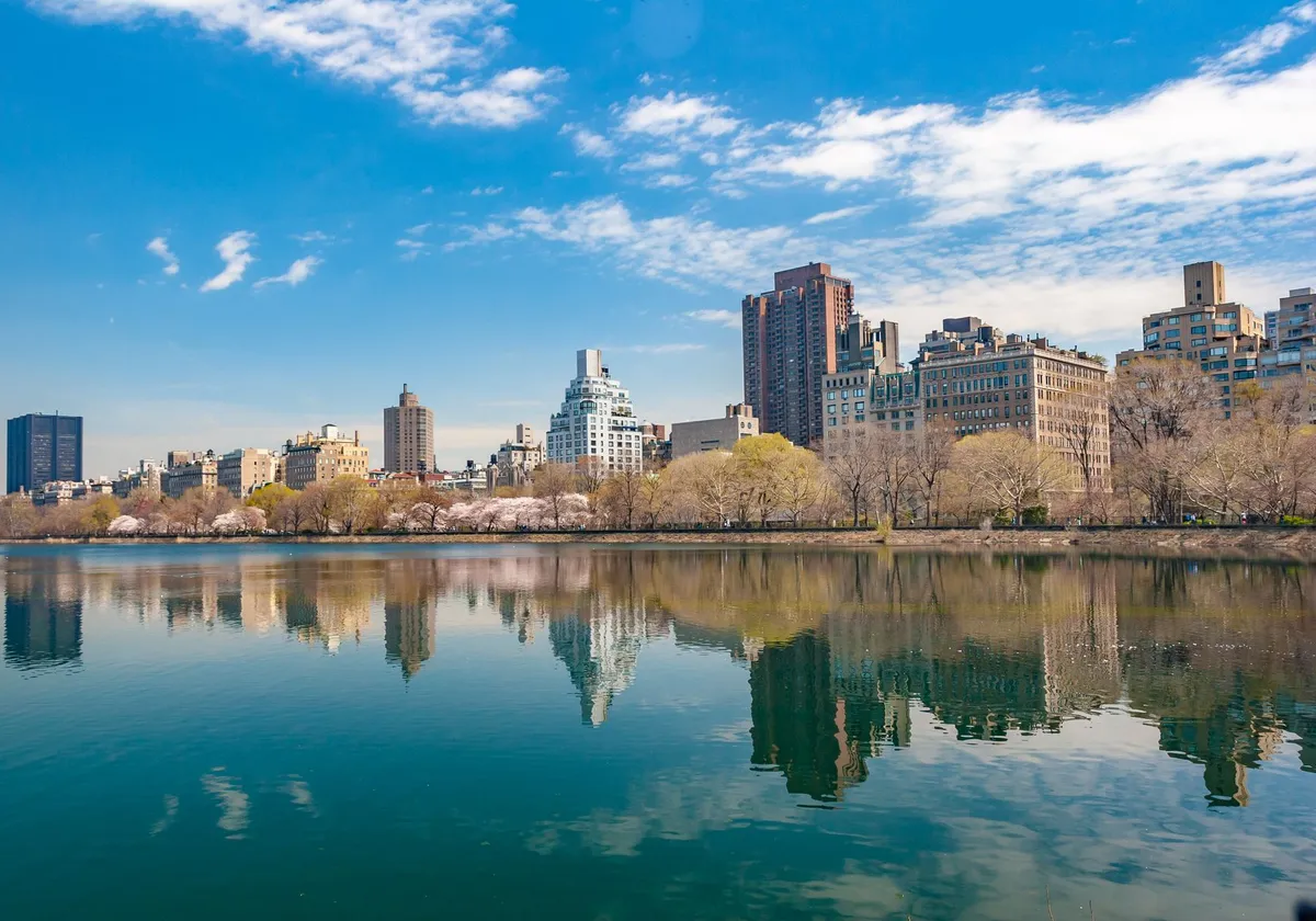 New York City skyline reflecting in Central Park's serene water under a vibrant blue sky.